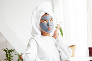 A woman in all white with a towel wrapped around her hair and mud mask on her face making a funny face.